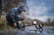 A person wearing a rain jacket kneels in the rain, actively using the Fisher Labs F44 The Ultimate Weatherproof Multi-Purpose Metal Detector, holding a small yellow object found during treasure hunting.