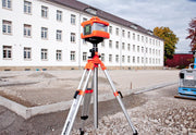 Nedo Heavy-Duty Elevating Tripod supporting a laser level on a gravel construction site in front of a white building.